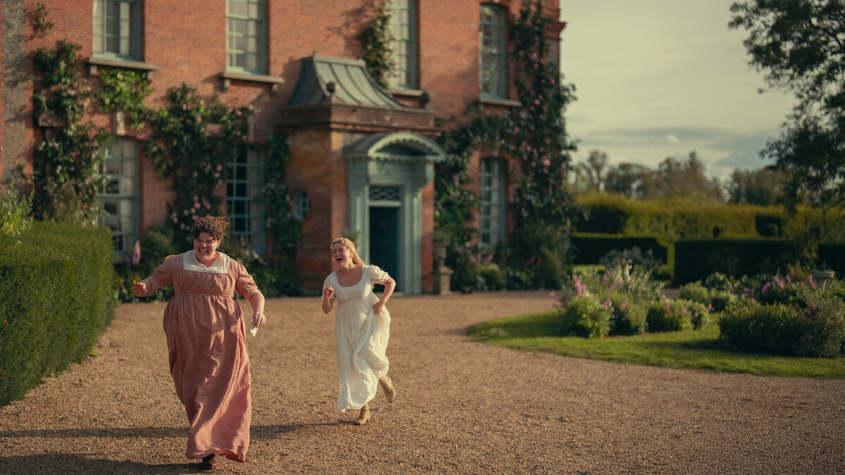 Two women in period dresses run joyfully on a gravel path in front of a large brick house with ivy and flowers, surrounded by hedges and gardens under a partly cloudy sky.
