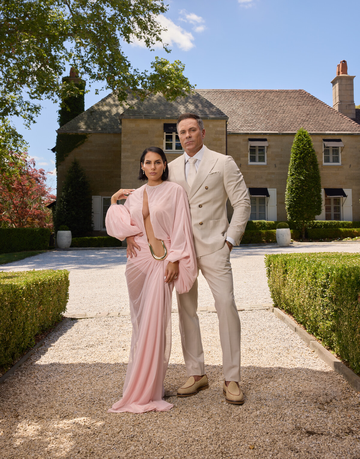 A well-dressed couple stands on a gravel path in front of a large, elegant house with manicured hedges and greenery on a sunny day. The woman wears a pink gown and the man wears a beige suit.