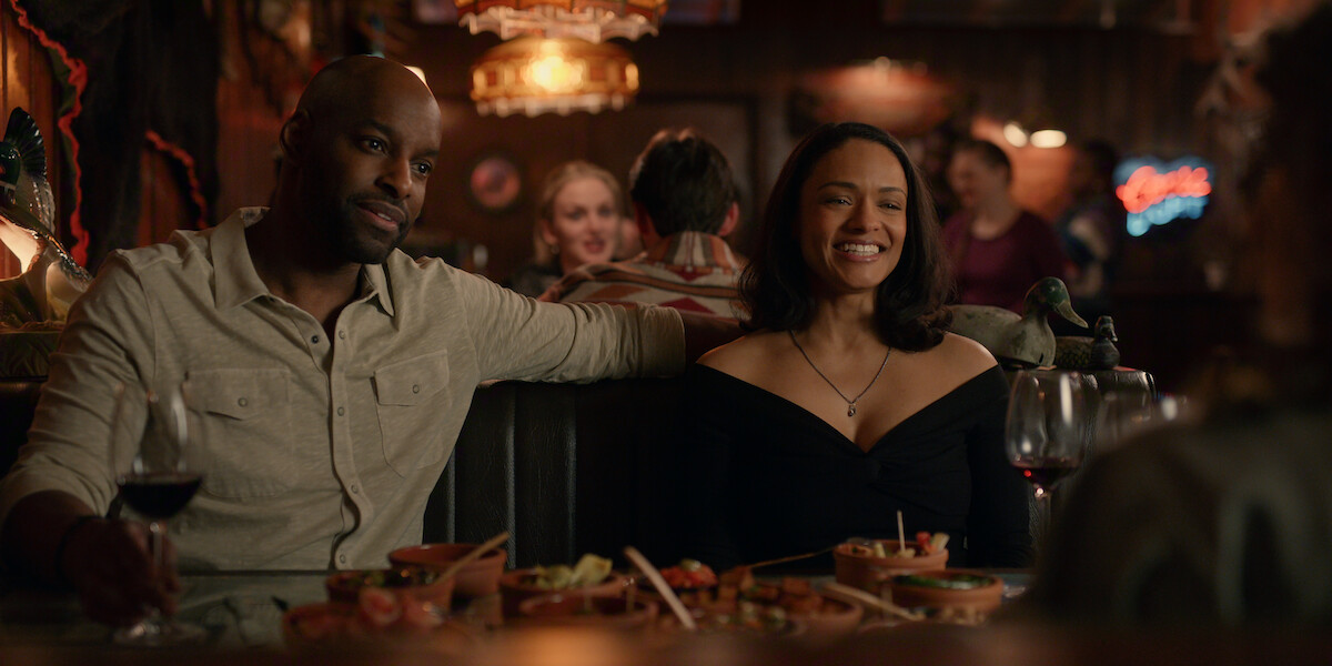 Two people sit together in a cozy restaurant booth, smiling and enjoying drinks and food, with a warm, inviting atmosphere and other diners in the background.