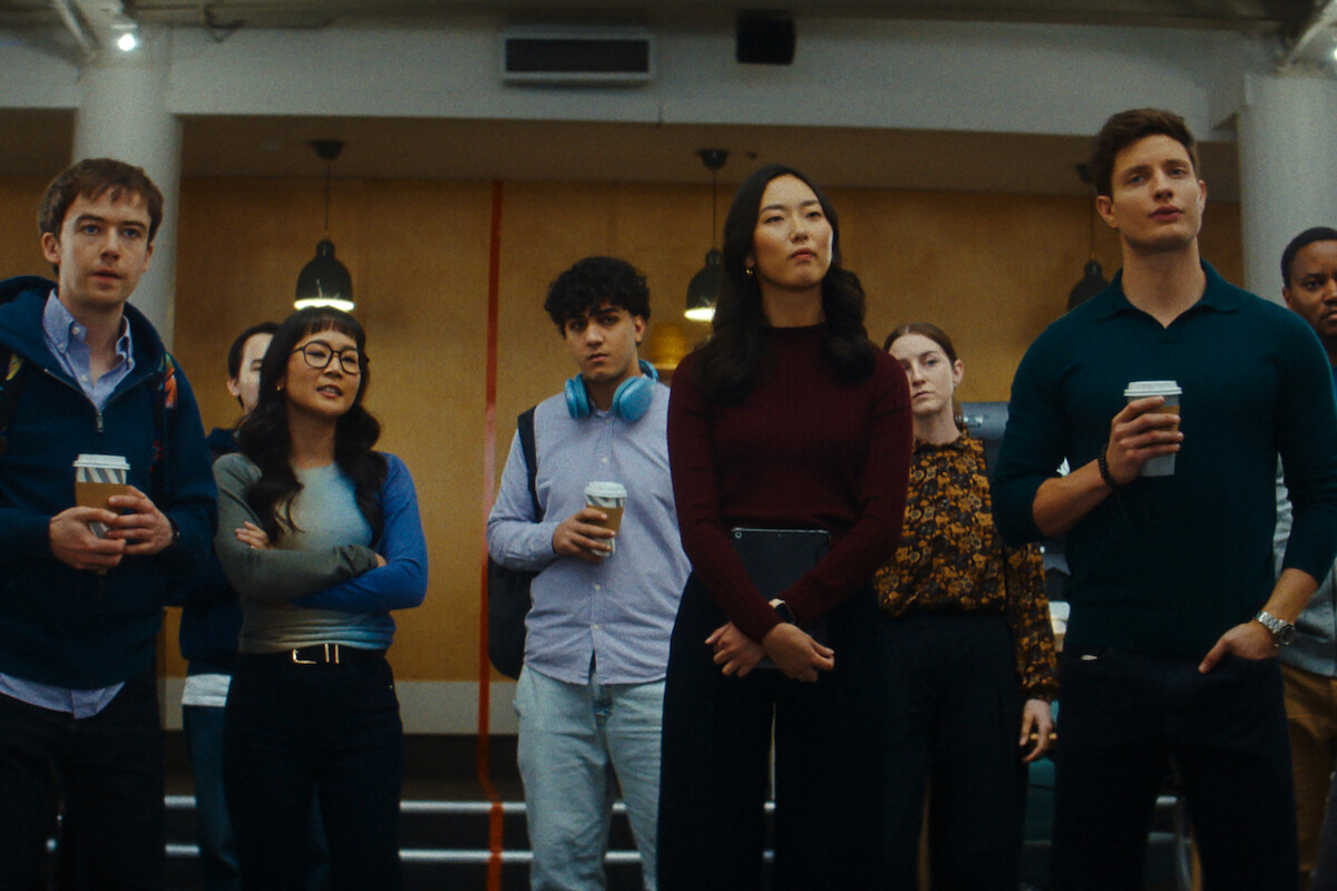 A group of young adults stands indoors in a modern office or meeting space, casually dressed, some holding coffee cups, looking attentively ahead under hanging lights.