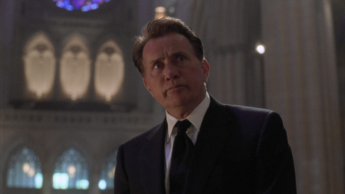 A man in a suit stands in a grand, dimly lit church or cathedral, looking upwards with a serious expression. The background features tall arches, stained glass windows, and dramatic lighting.