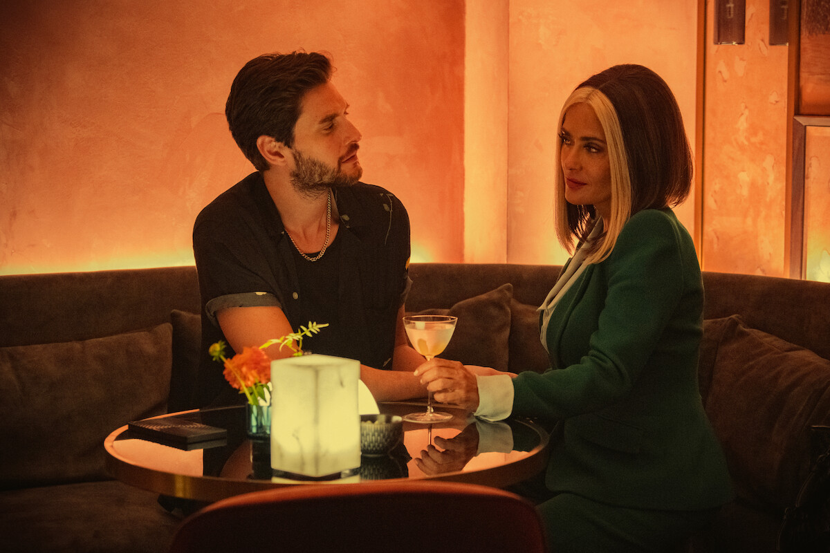 Ben Barnes as TV Mac and Salma Hayek as TV Joan in Black Mirror Season 6 sitting at a table sharing a cocktail.
