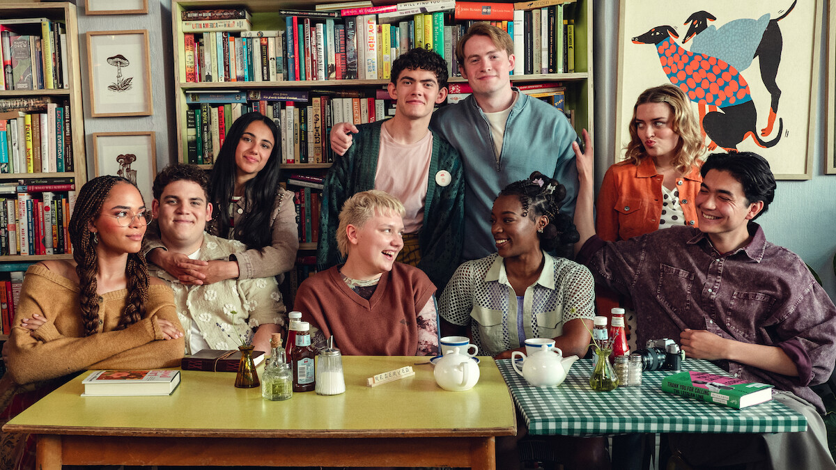 A group of eight young people sit and stand together around a table with tea and books in a cozy, colorful room lined with bookshelves and decorated art, showing warmth and friendship.