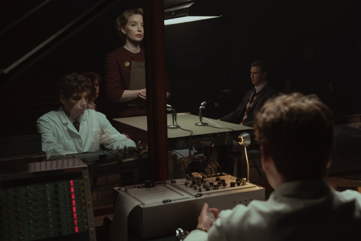 A group of people in a dimly lit room, some in lab coats, appear to be conducting or observing an experiment or interrogation through glass, with vintage recording equipment and microphones on the desk.