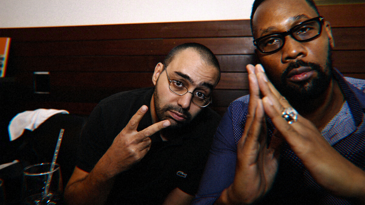 Two men sitting in a dimly lit indoor setting with wooden paneled walls, one making a peace sign and the other with hands clasped, both wearing glasses and casual shirts.