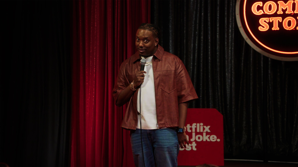 Comedian performing stand-up on stage, holding a microphone. Red and black curtains, a neon "Comedy Store" sign, and a "Netflix is a Joke Fest" sign in the background. Indoor comedy club environment.