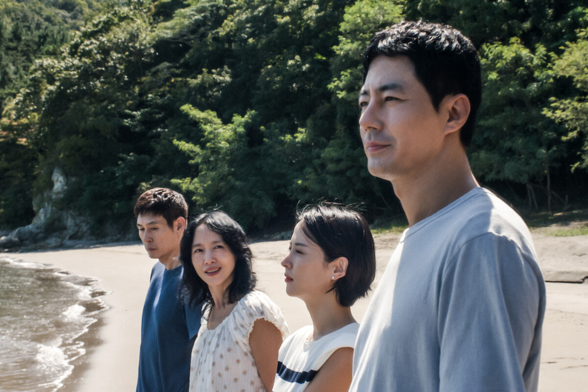 Four people stand side by side on a sandy beach, facing the water, with green trees and sunlight in the background. They appear thoughtful and relaxed, enjoying a peaceful day at the shoreline.