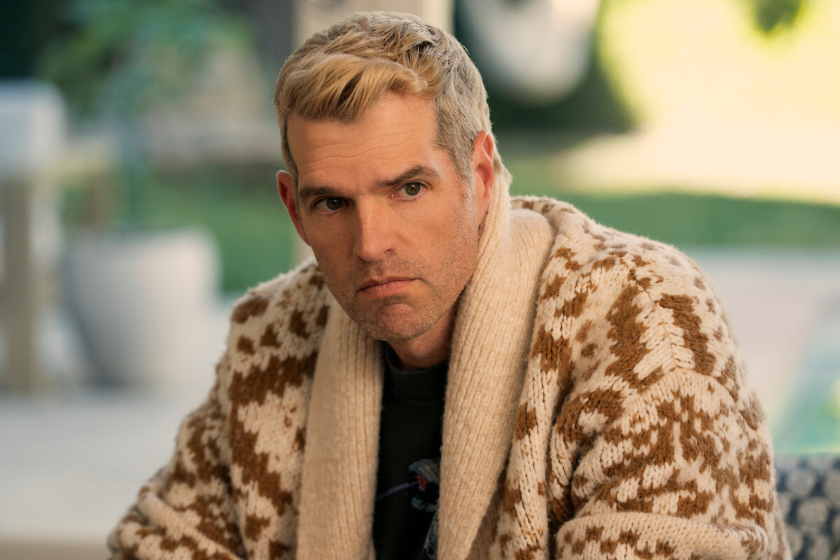 A man with short blonde hair wearing a patterned knit cardigan sits indoors, looking serious. The background is softly blurred, suggesting a bright room with green outdoor scenery visible through windows.