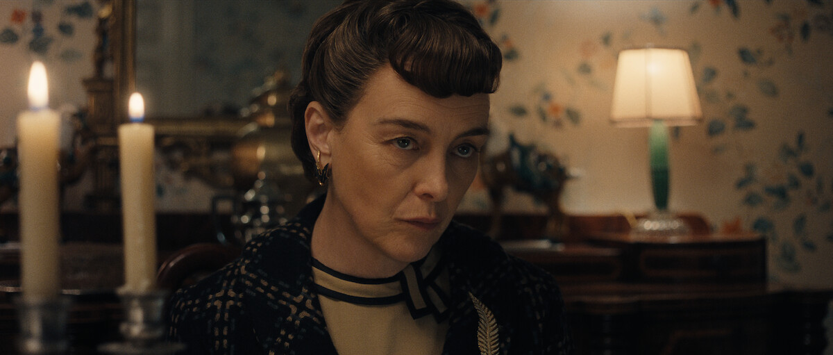 A woman with short, styled hair and a serious expression sits at a table with two lit candles, in a room decorated with floral wallpaper and a lamp in the background.