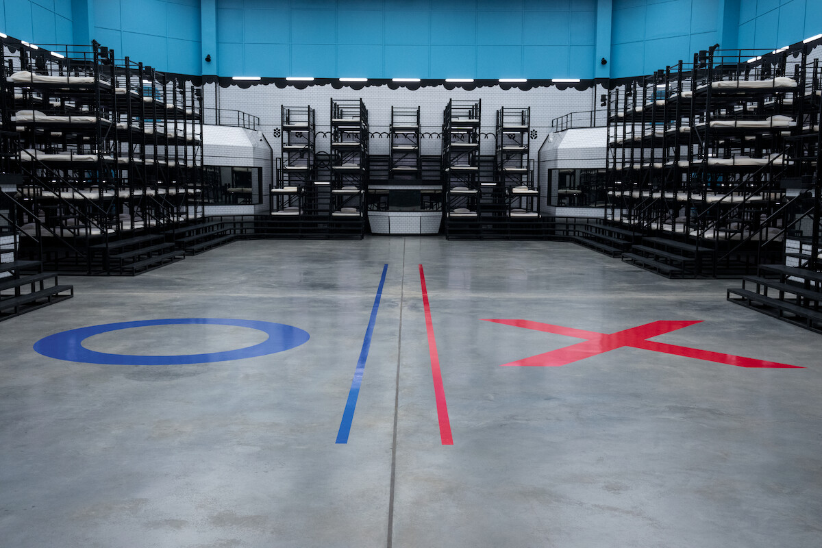 Empty, modern indoor arena with arranged black seating platforms, blue "O" and red "X" symbols marked on polished concrete floor, stark lighting, futuristic mood, possibly set for a competition or event.