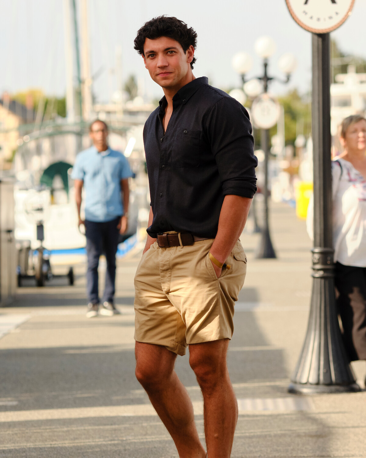 Man in black shirt and khaki shorts stands with hands in pockets on a marina dock; boats, people, and street lamps in background, sunny day.