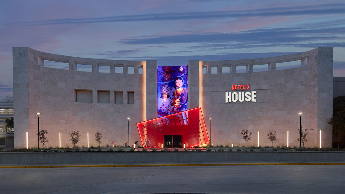 Modern stone building at dusk with a digital screen showing animated characters, bold red “Netflix House” sign, vibrant lighting, and a welcoming entrance, creating a lively and entertaining atmosphere.