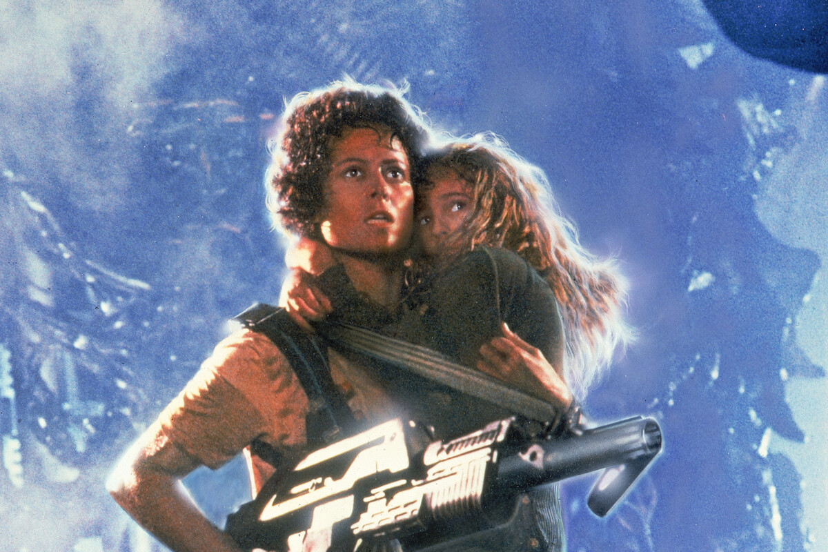 A woman holding a large futuristic gun stands protectively in a smoky, alien environment with a scared young girl clinging to her, both looking alert and tense amidst a dramatic, sci-fi backdrop.