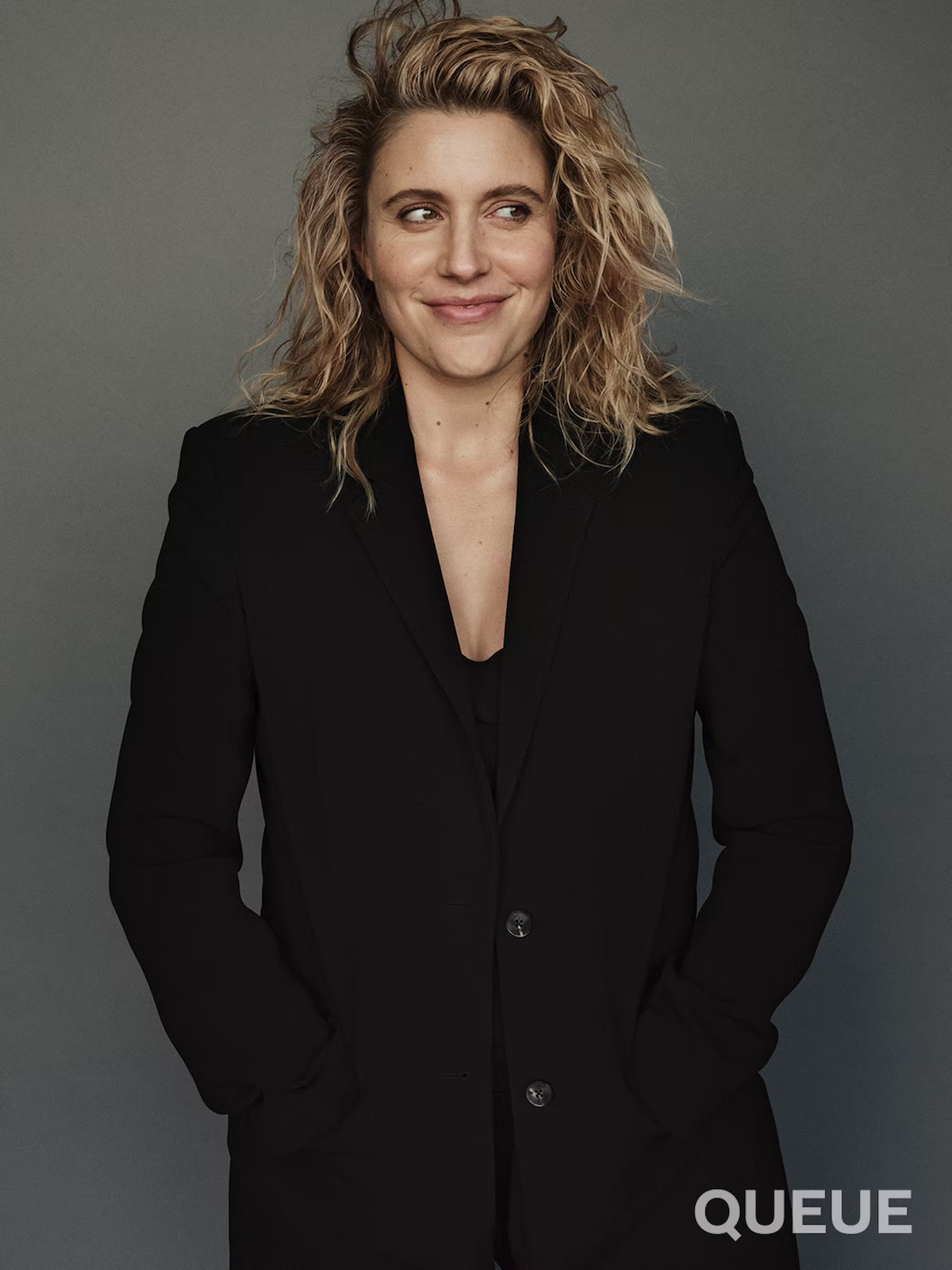 Greta Gerwig wears a black jacket against a gray backdrop.