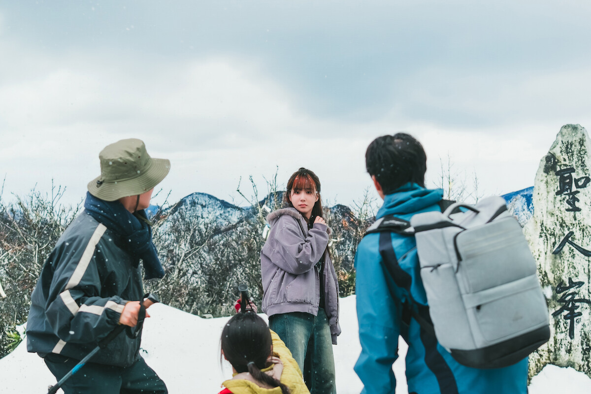 Four people, including a woman in a purple jacket, stand on a snowy mountain with hiking gear. They are outdoors near a stone marker with Japanese writing, surrounded by winter trees and cloudy skies.