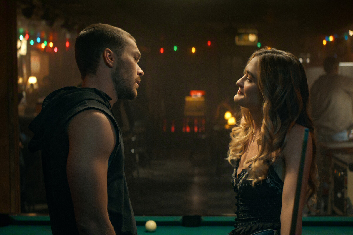 A man and woman stand close by a pool table in a dimly lit bar with string lights, facing each other and engaged in conversation, background out of focus and atmospheric.