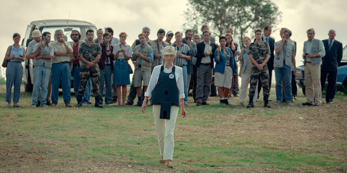 A crowd of people standing together outside in the background looking at a Diana walking away in the foreground.