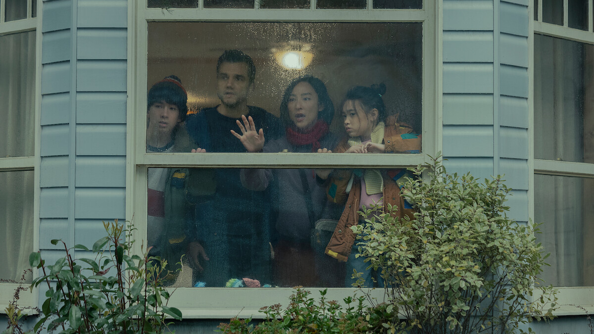 Four people stand closely together indoors, looking anxiously out a rainy window of a house with pale siding, surrounded by green shrubs, under soft indoor lighting.
