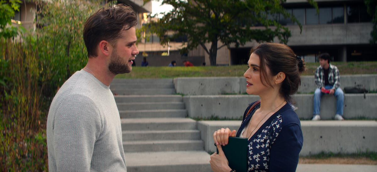 A man and woman are talking outside on concrete steps on a campus; she holds a folder, with another person sitting in the background. Trees and a building are visible.