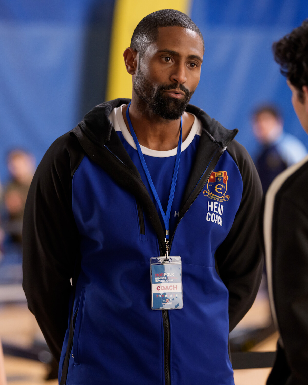 A basketball head coach wearing a team jacket and lanyard badge stands indoors in a gymnasium, talking to a player. The environment includes blurred gym equipment and people in the background.