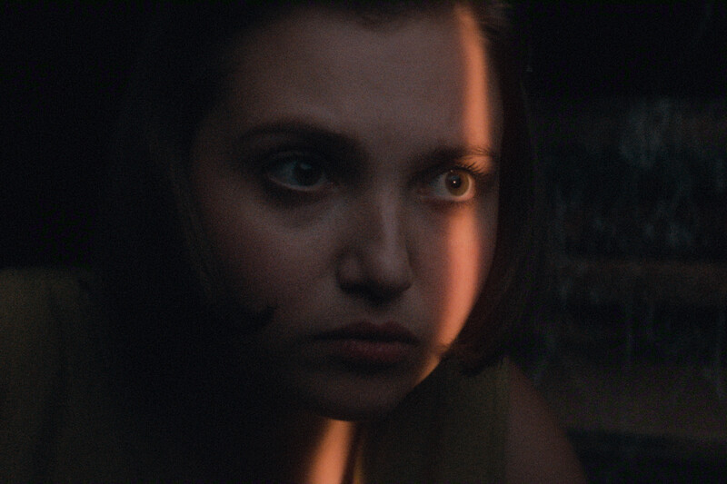A young woman with short hair sits in a dimly lit room, a narrow ray of light illuminating part of her face. She appears tense or worried, with a serious expression, creating a suspenseful and dramatic mood.