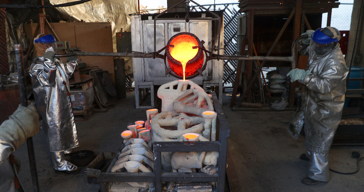 two people in high temperature suits pour molten steel into a cast