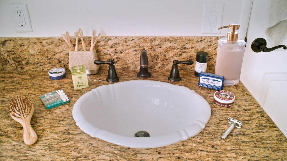 Bathroom sink with brown granite counter, flanked by personal care items like toothbrushes, soap, brush, razor, toothpaste, lotion, and other toiletries arranged around the white basin in a well-lit, tidy bathroom setting.