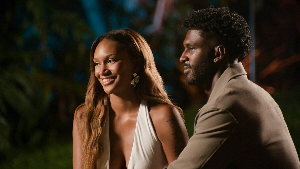 A smiling woman in a white dress sits beside a man in a tan jacket at night, outdoors with greenery and colorful lights in the background.