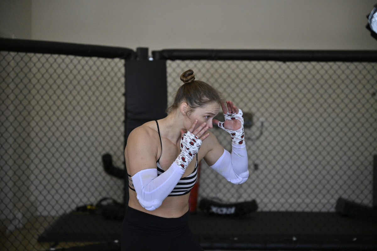 Ronda Rousey and Kehlani laughing together in an MMA training gym, standing on mats near a fenced cage, wearing athletic workout clothing with one sporting visible tattoos.