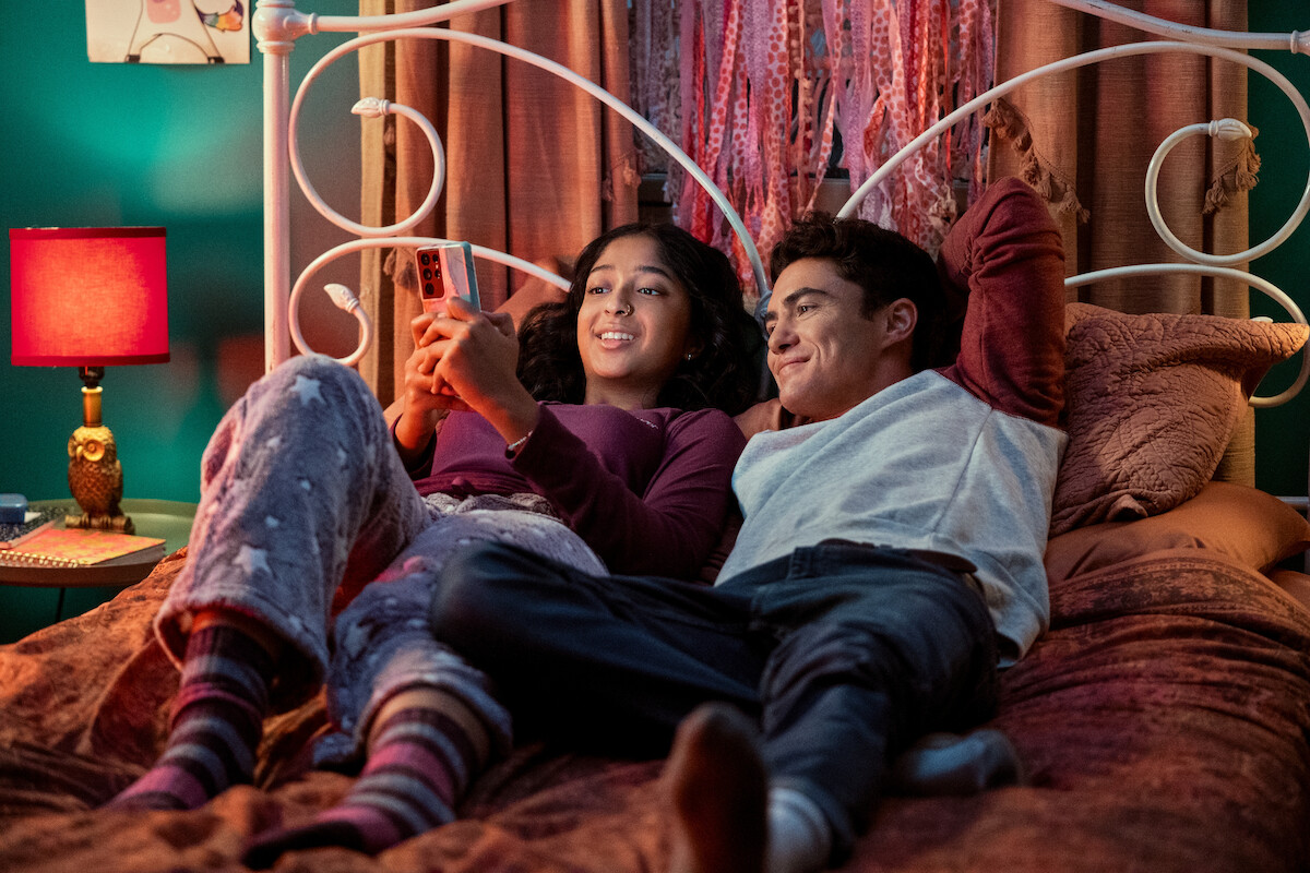 Maitreyi Ramakrishnan as Devi in Never Have I Ever Season 1 and Darren Barnet as Paxton Hall-Yoshida in Never Have I Ever Season 3 laying down on bed looking at phone.