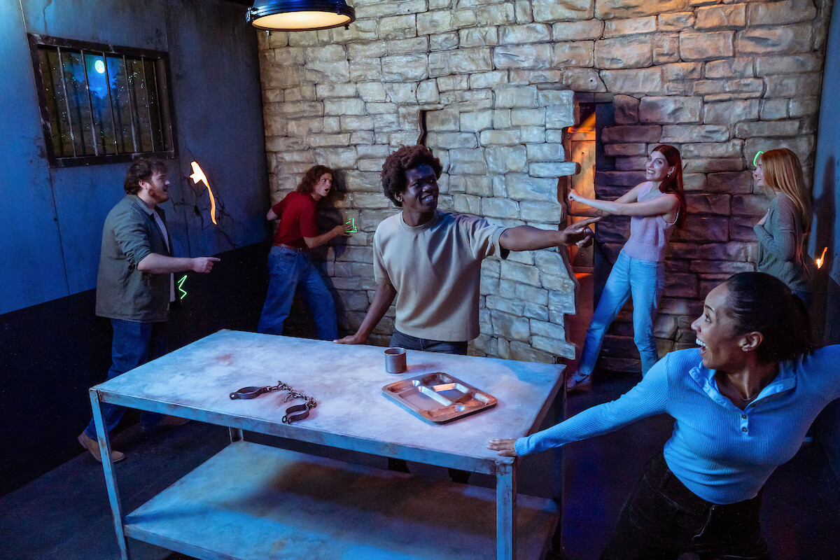 Group of people solving puzzles in an escape room with stone walls, a metal table, and various props, working together enthusiastically to unlock a secret door in a dimly lit environment.
