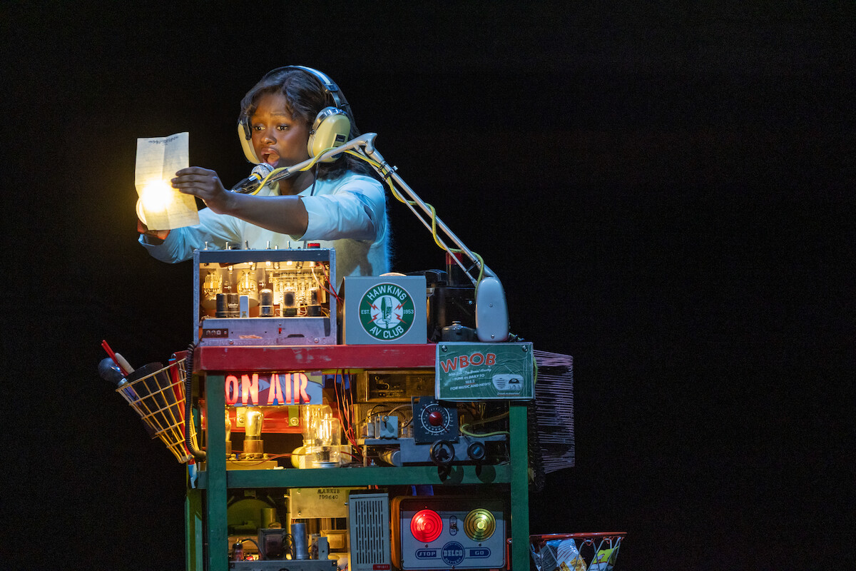 Person wearing headphones sits at a colorful, gadget-filled broadcast desk on a dark stage, speaking into a microphone and holding a sheet of paper illuminated by a bulb, surrounded by tech equipment, lights, and a Starbucks logo.