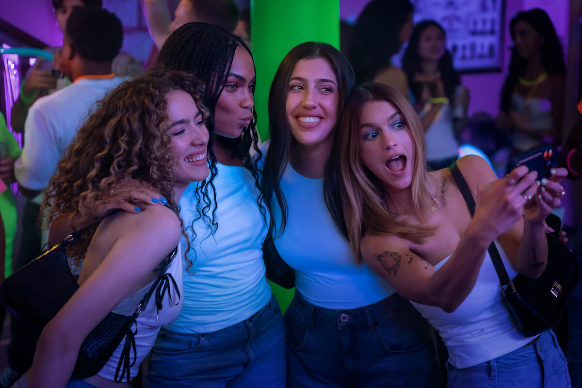 Four young women in casual clothes take a selfie together at a lively indoor party with colorful neon lighting and other people in the background.