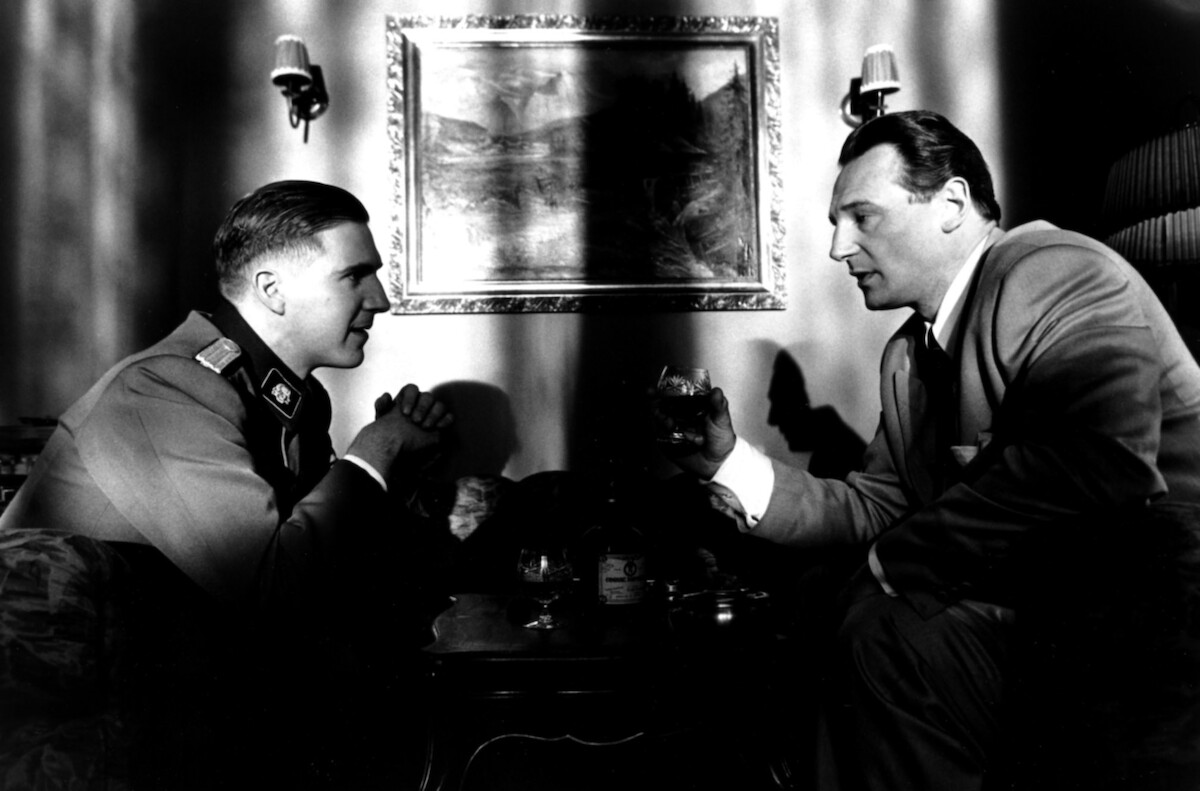 Ralph Fiennes and Liam Neeson sit across from each other at a table in ‘Schindler’s List’
