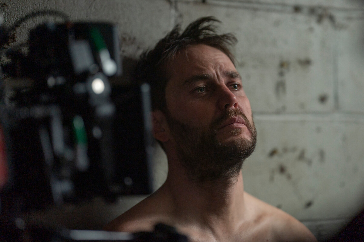 Taylor Kitsch as Glen Kryger leans his head against a cement wall as a camera films him in ‘Painkiller.’