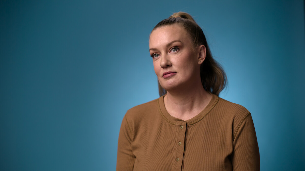 Woman with light hair in a ponytail wearing a brown button-up shirt sits in front of a blue background, looking slightly off camera with a thoughtful expression in a studio setting.