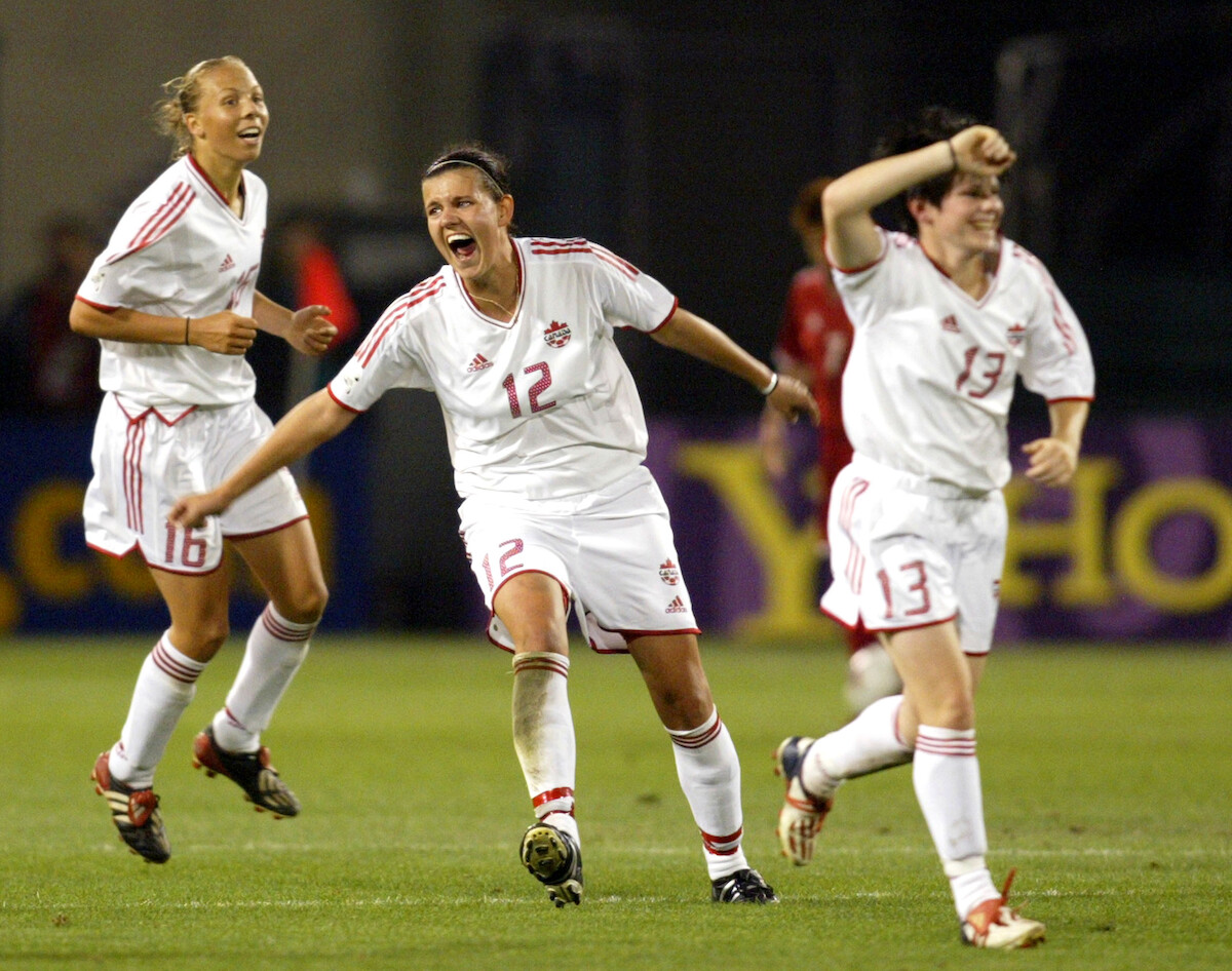 Three female soccer players in white uniforms celebrate energetically on a green field during a night match, with a blurred stadium background and other players in the distance.