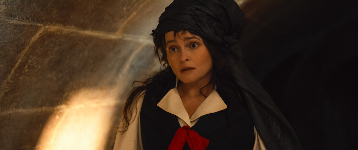Woman dressed in period costume with a black headdress and red ribbon stands in a dimly lit stone hallway, looking surprised or concerned. Warm light highlights the stone wall in the background.