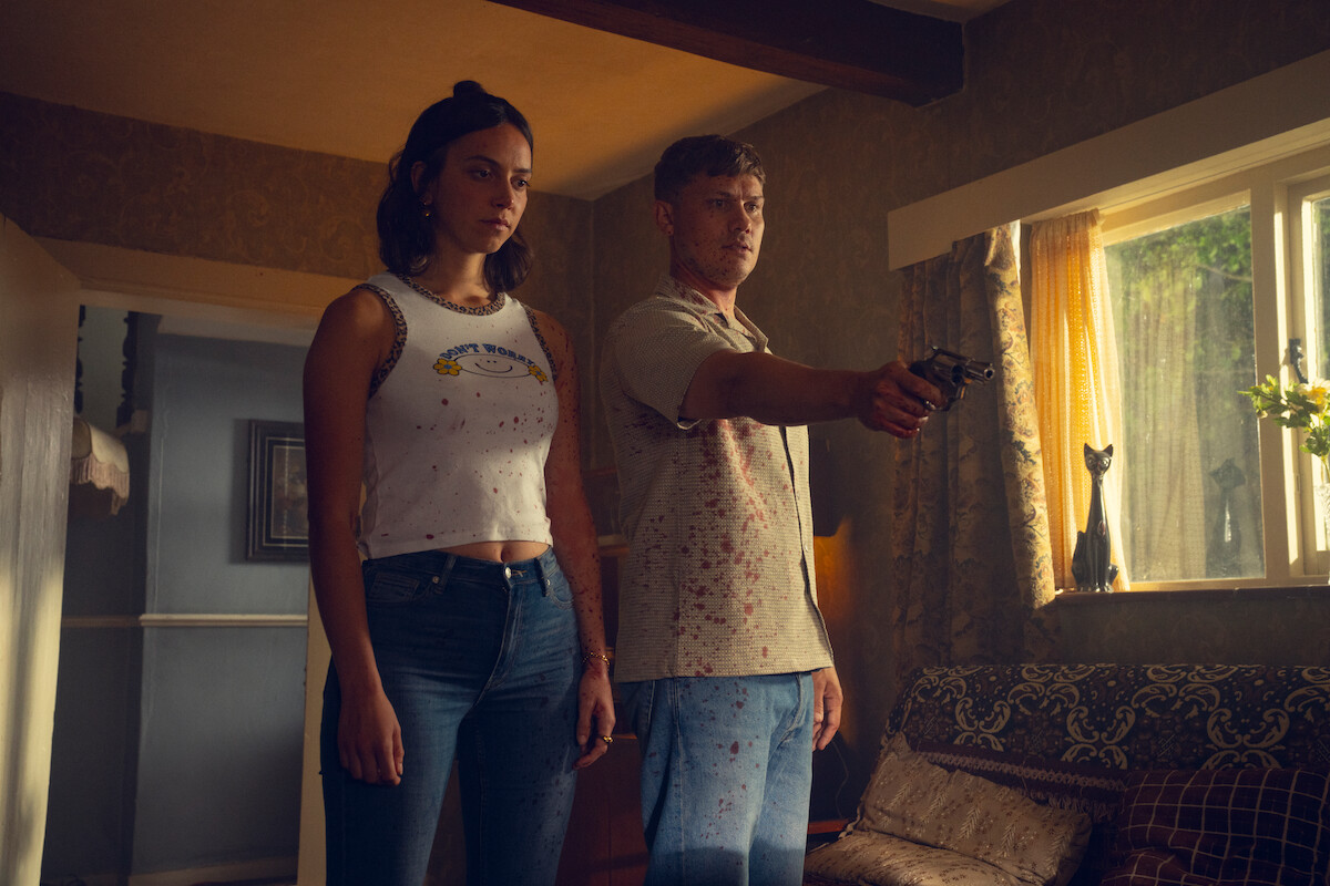 A man aiming a gun and a woman standing beside him in a dimly lit living room with sunlight streaming through the window, creating a tense and dramatic atmosphere.