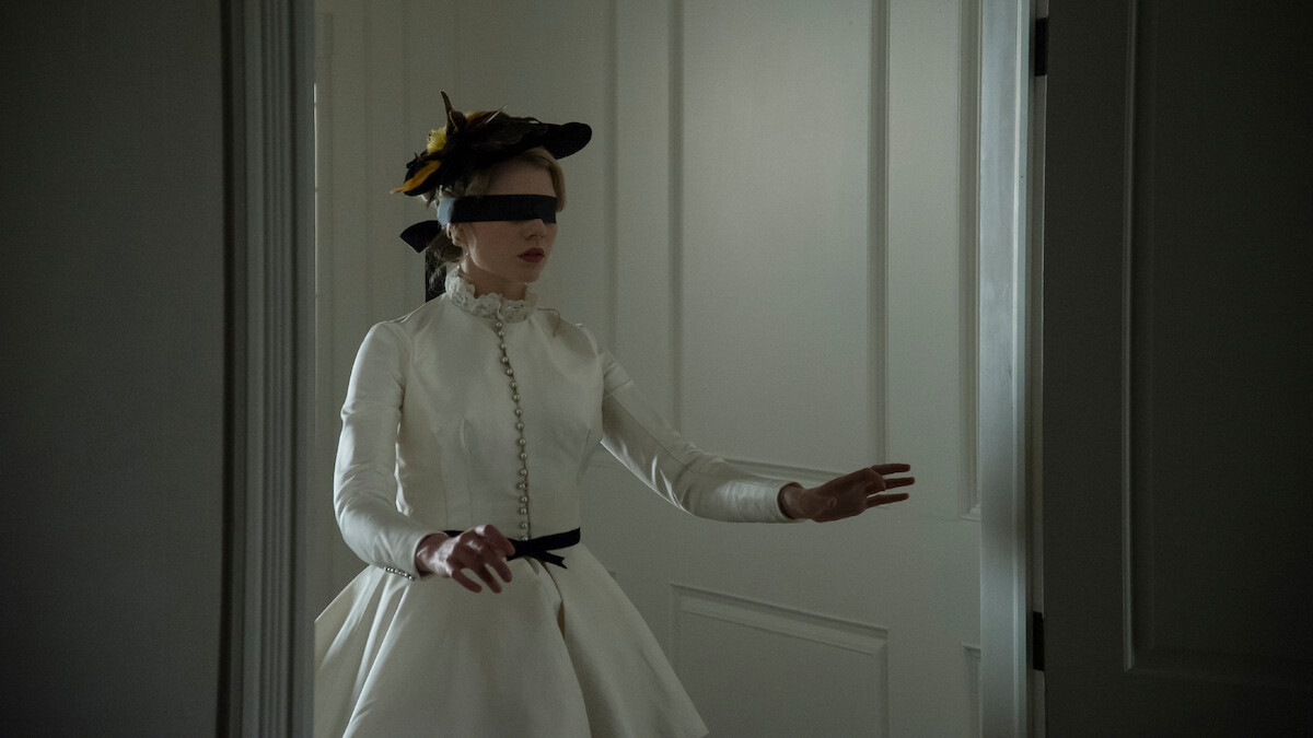 A woman in a white period costume and feathered hat, blindfolded, cautiously steps forward with arms outstretched inside a dimly lit room with white paneled doors.