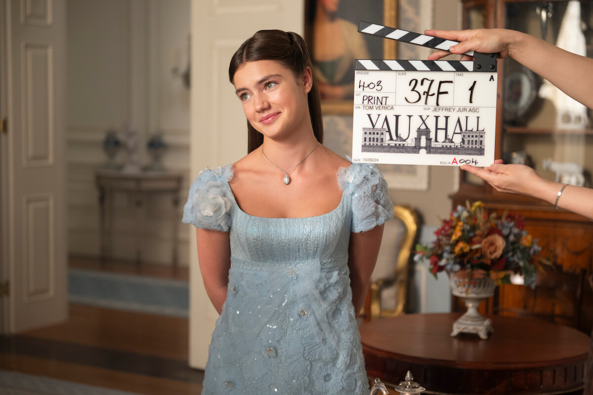 Uma jovem com um vestido de época azul-claro está de pé em uma sala decorada com flores e móveis clássicos, enquanto alguém segura uma claquete à sua frente, indicando a gravação de uma cena.