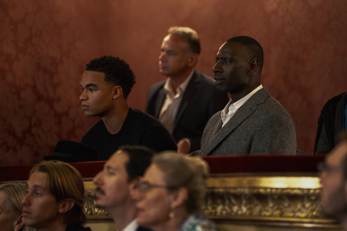 Dois homens negros aparecem em primeiro plano em uma tribuna de teatro, rodeados por outras pessoas