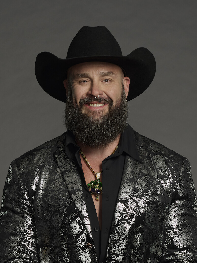 Homem com barba usando um chapéu de caubói preto, blazer preto com estampas e um colar, posa em frente a um plano de fundo escuro, sorrindo para a câmera em um ambiente de estúdio.