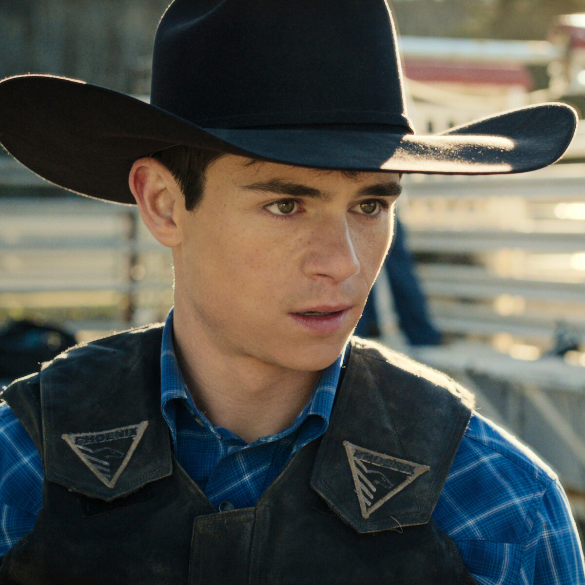Young man in cowboy hat and plaid shirt stands outdoors at a rodeo or ranch setting, wearing a protective vest, talking to another cowboy with livestock and fencing in the background.