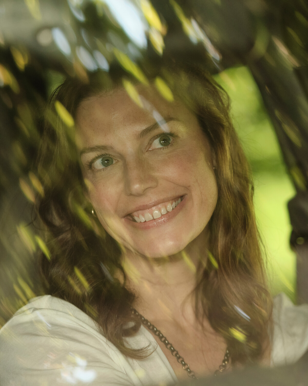 Smiling woman with wavy hair sits inside a car, viewed through the window with reflections of green trees and natural light outside, creating a soft, serene atmosphere.