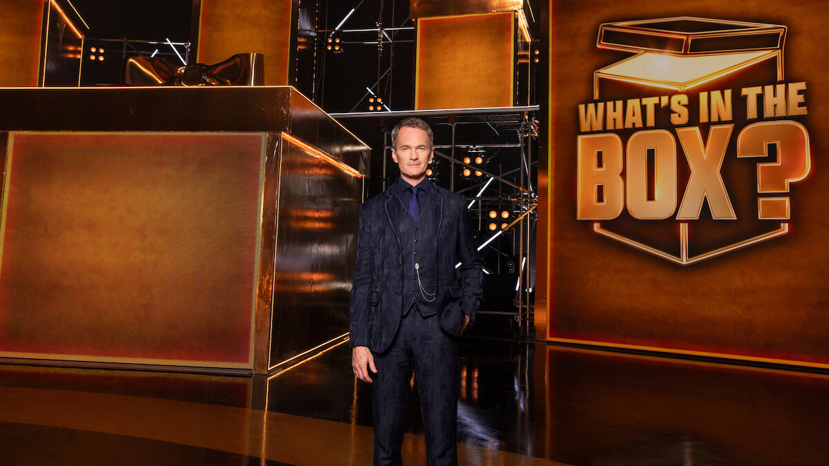 A man in a dark suit stands on a shiny stage with large golden boxes and a big sign reading "WHAT'S IN THE BOX?" The environment is dramatic with glowing lights and a game show atmosphere. Mood is suspenseful and exciting.