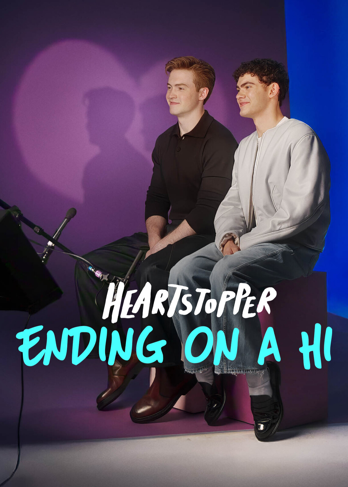 Two young men sit side by side on a purple bench in a studio with purple and blue lighting, microphones in front, smiling, with Heartstopper: Ending on a Hi text overlayed.