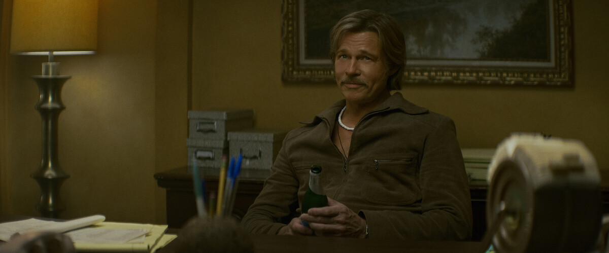 A man with a mustache sits at a desk in a warmly lit office, holding a green bottle. The room has a lamp, filing boxes, and papers, with a painting and organized supplies in the background.