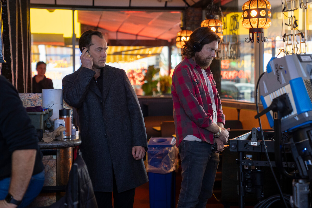 Two men stand in a diner filled with film equipment and lighting; one wears a dark coat, the other a plaid shirt, both appear focused, suggesting a movie or TV production setting with a casual, behind-the-scenes atmosphere.