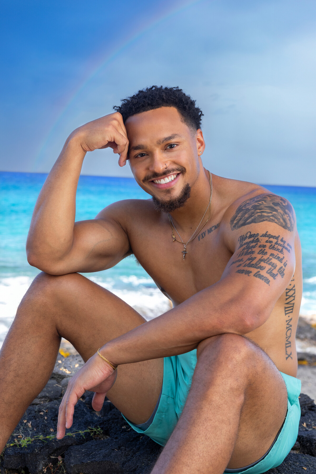 Smiling shirtless man with tattoos sits on rocks by turquoise ocean under a blue sky with a rainbow, wearing aqua swim trunks, relaxed and enjoying the beach environment.
